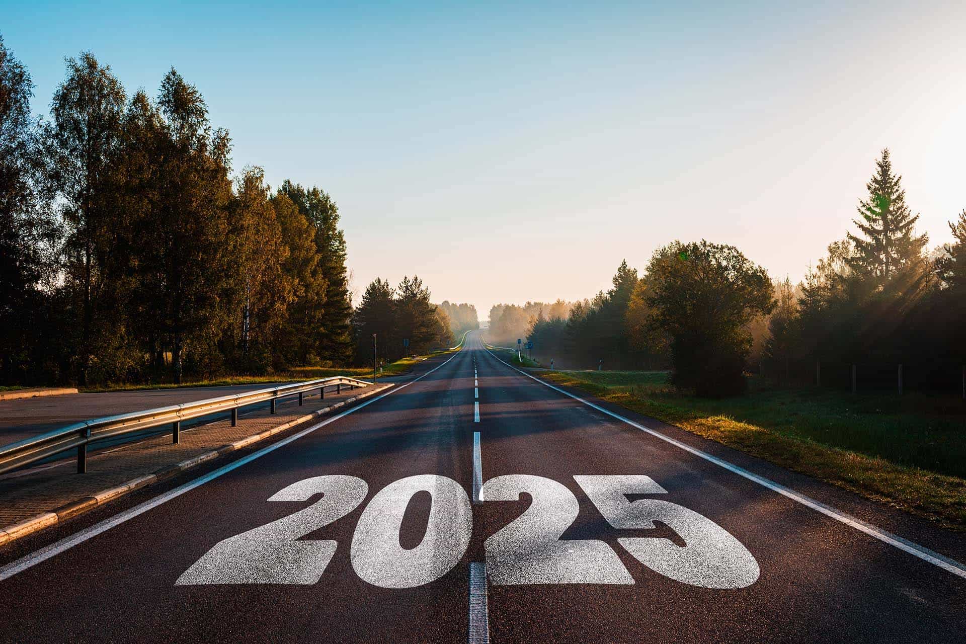 A two-lane road with "2025" painted in bold white numbers leads into the distance, surrounded by lush greenery and illuminated by a warm sunrise, evoking themes of progress and new beginnings.